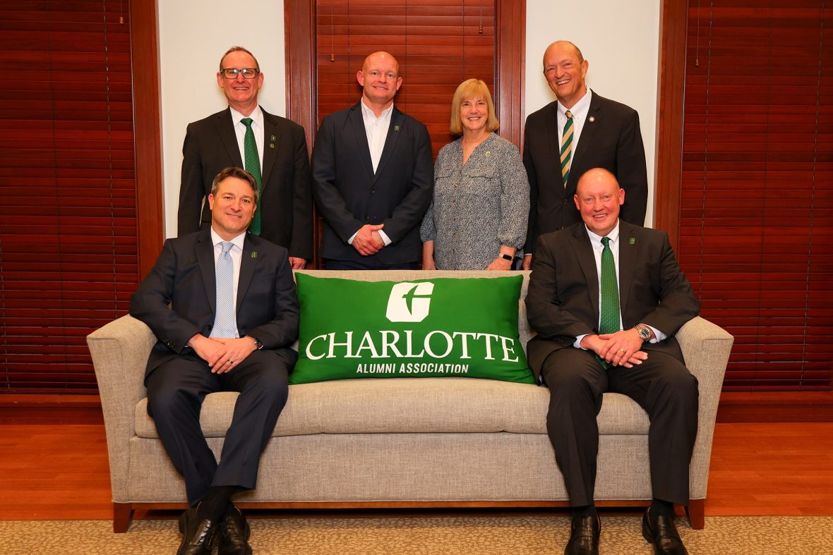 Two people sit on opposite ends of a couch with a UNC Charlotte Alumni Association green pillow between them, and four people stand behind them