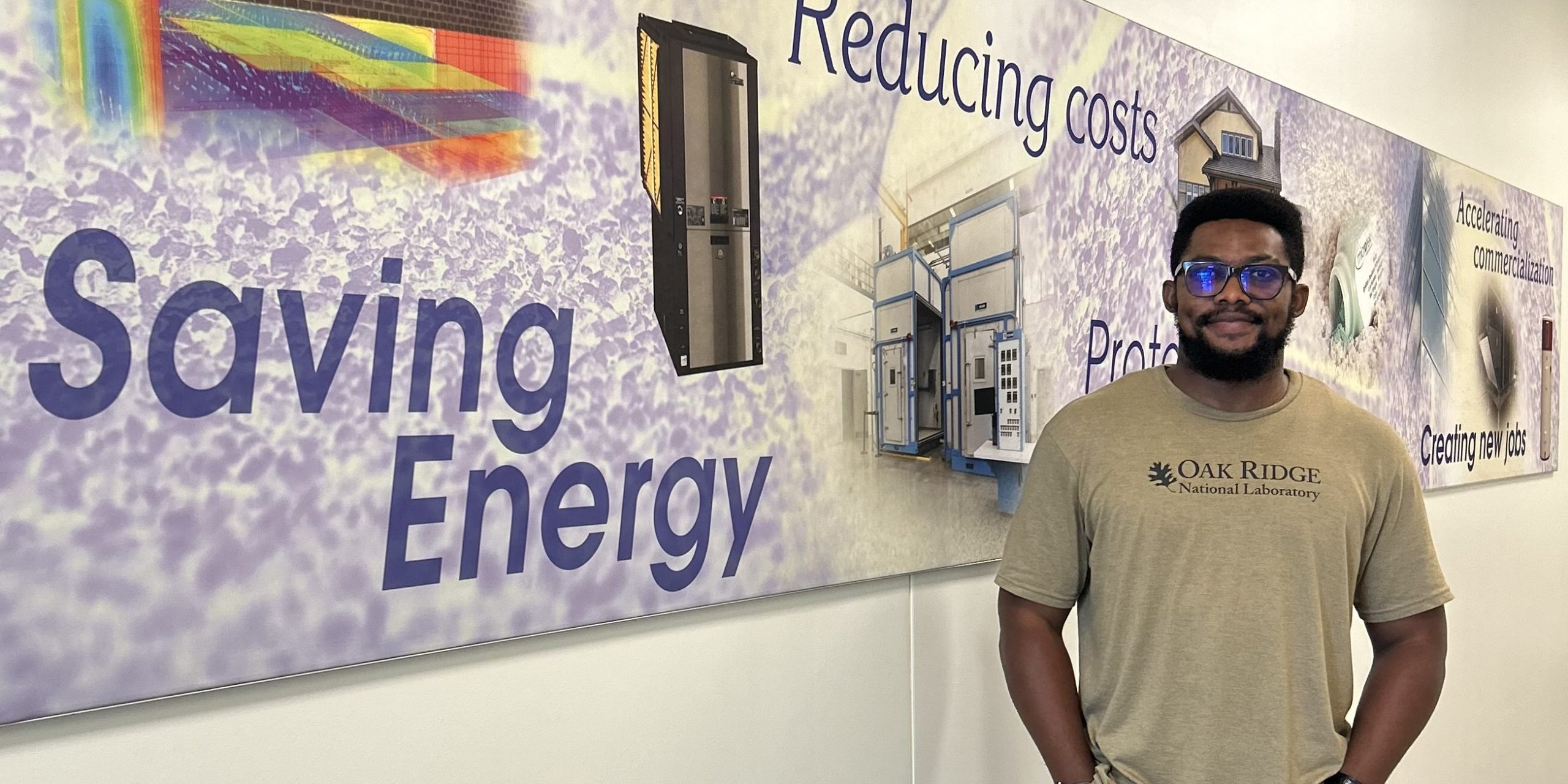 Graduate student Felix Ekuful stands in front of a panel with text reading "saving energy," "reducing costs," "accelerating commercialization" and "creating new jobs."