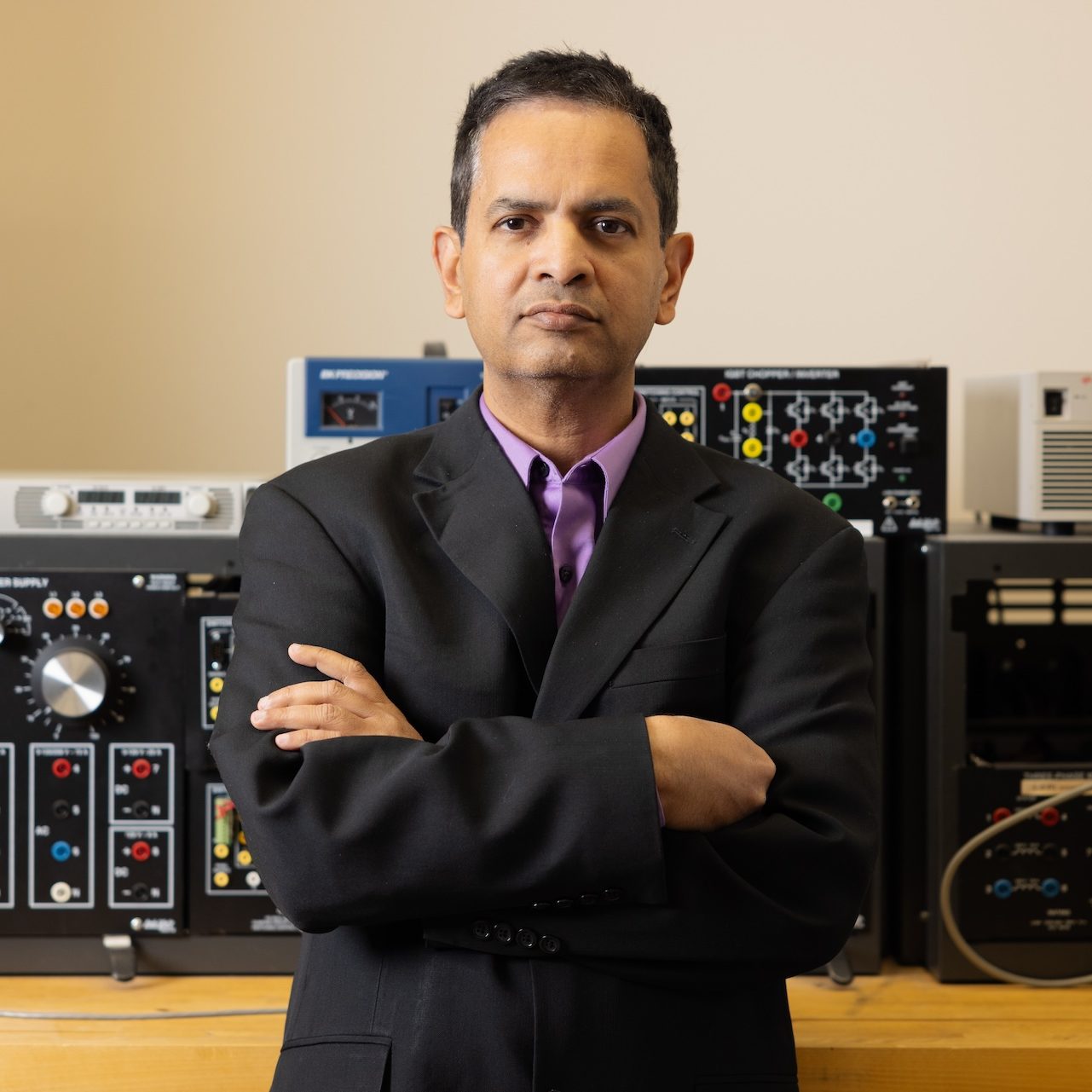 Portrait of Sukumar Kamalsadan, wearing a suit with his arms crossed, while standing in front of equipment 