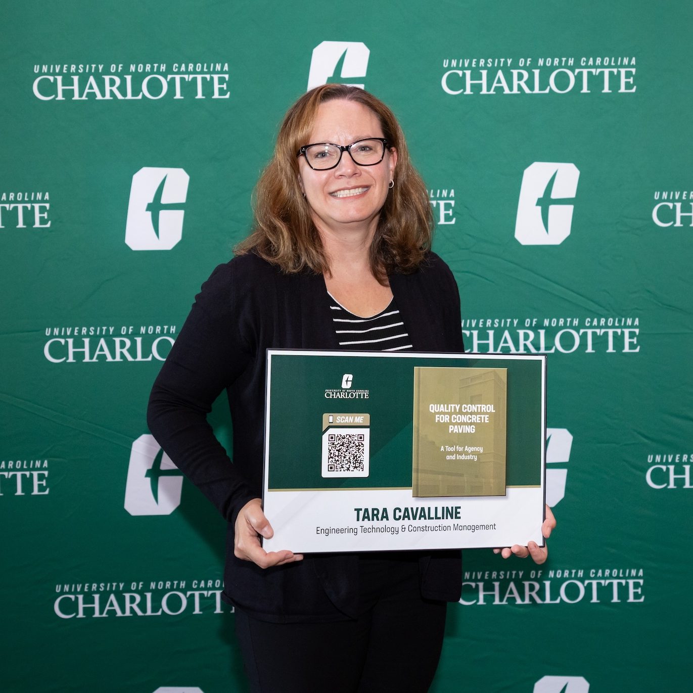 Tara Cavalline holds a poster while standing in front of a green background with the white Charlotte logo repeated on it