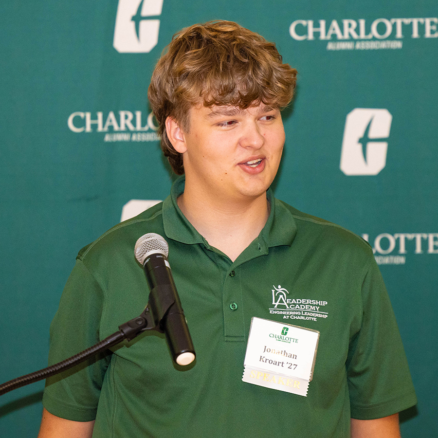 Jonathan Kroart speaks at a podium in front of a green backdrop featuring the Charlotte Alumni Association logo.