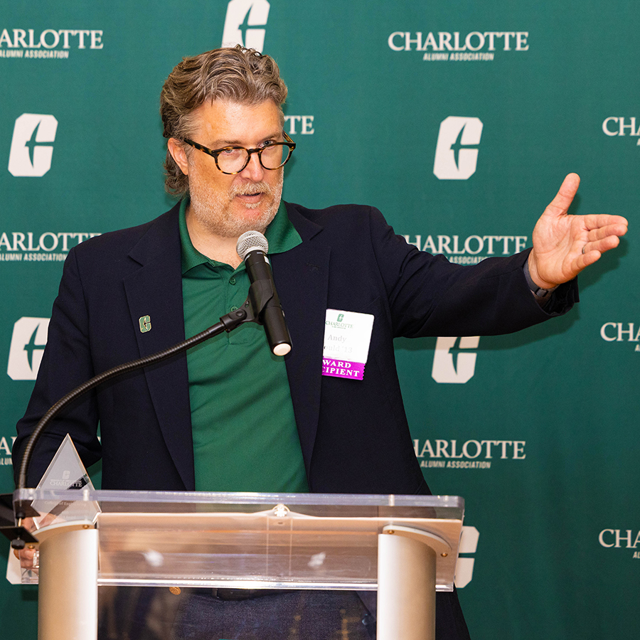 Andy Gould speaks at a podium in front of a green backdrop featuring the Charlotte Alumni Association logo.