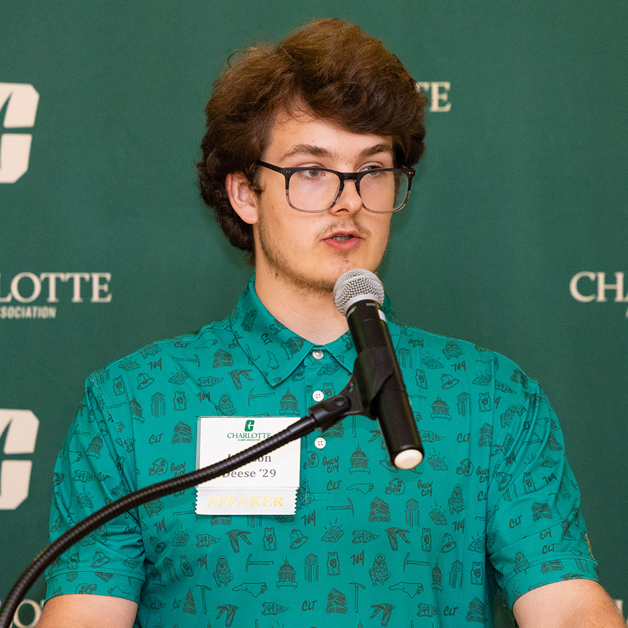 Jackson Deese with glasses stands at a podium speaking into a microphone in front of a green backdrop featuring the Charlotte Alumni Association logo.