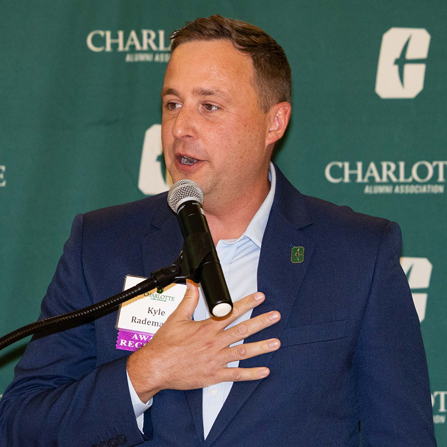 Kyle Rademaker in a suit stands at a podium speaking into a microphone during an event hosted by the Charlotte Alumni Association.