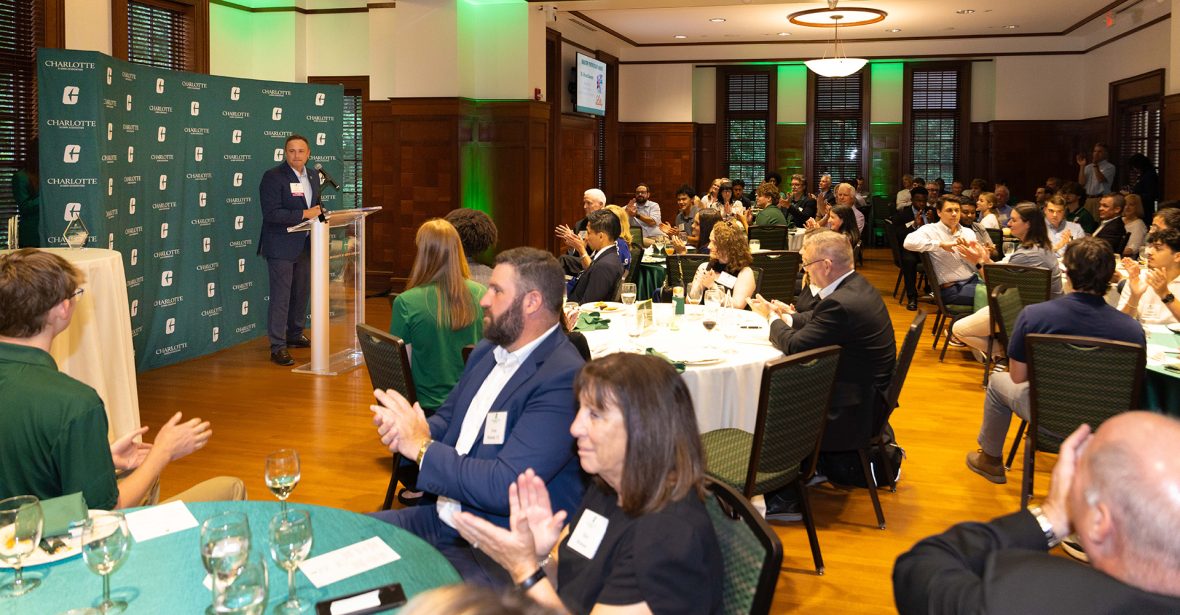 A speaker addresses an audience at a formal event in a banquet hall, with attendees seated at round tables and applauding.