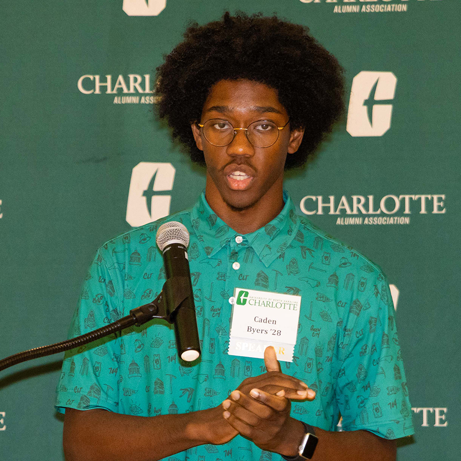 Caden Byers stands at a podium speaking into a microphone during an event hosted by the University of North Carolina Charlotte Alumni Association.