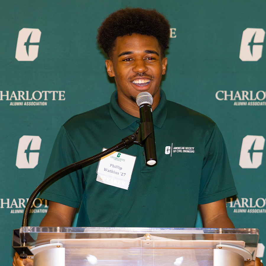 Phillip Watkins stands at a podium speaking into a microphone in front of a backdrop featuring the Charlotte Alumni Association logo.