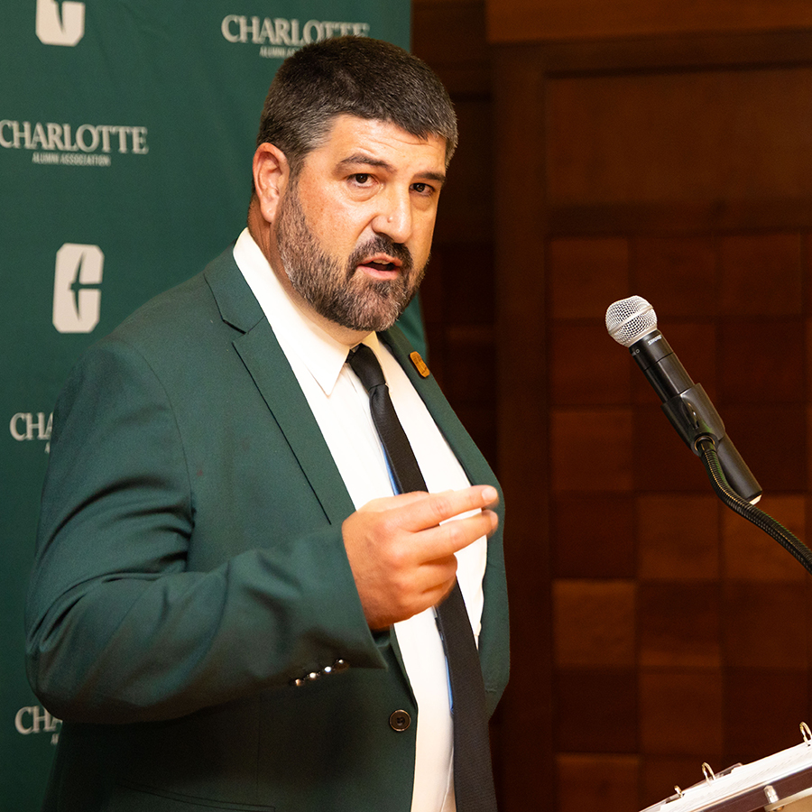 Ashton Watson in a dark suit stands at a podium speaking into a microphone, with a backdrop featuring the Charlotte Alumni Association logo.