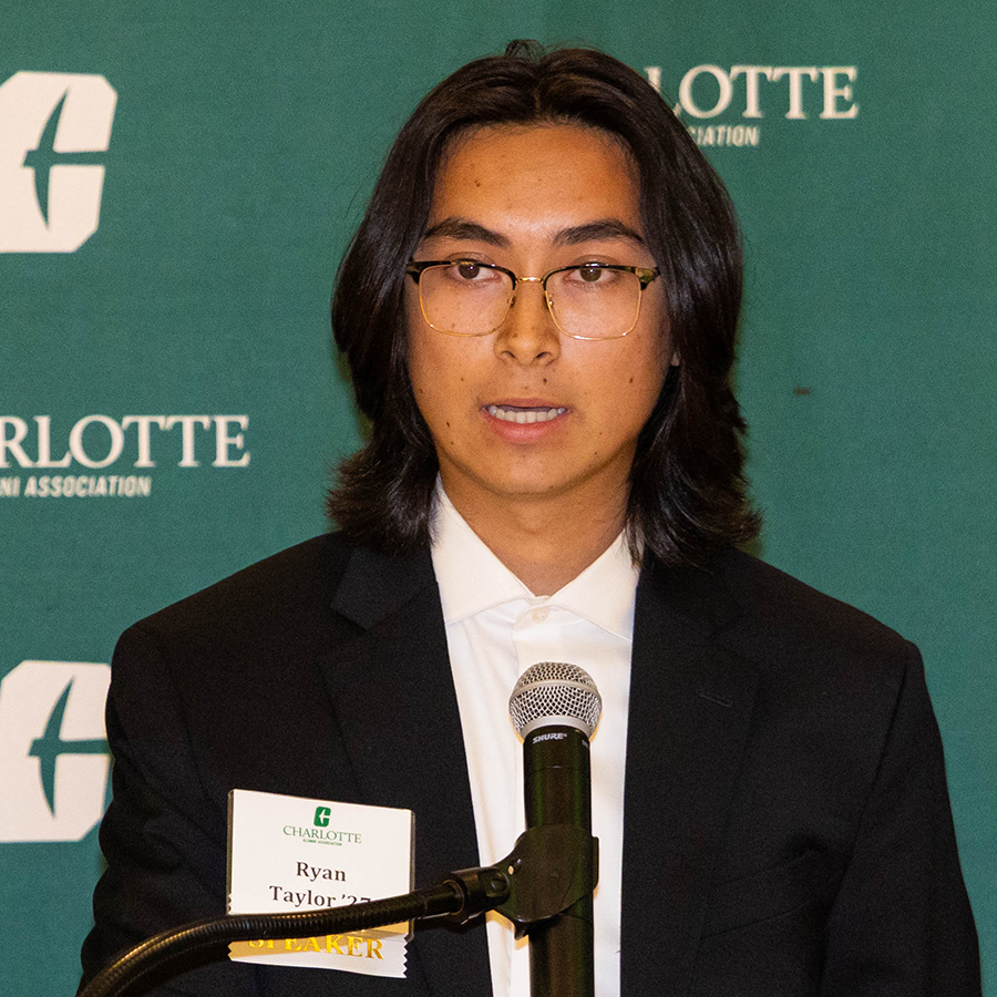 Ryan Taylor stands at a podium speaking into a microphone in front of a green backdrop featuring the Charlotte Alumni Association logo.