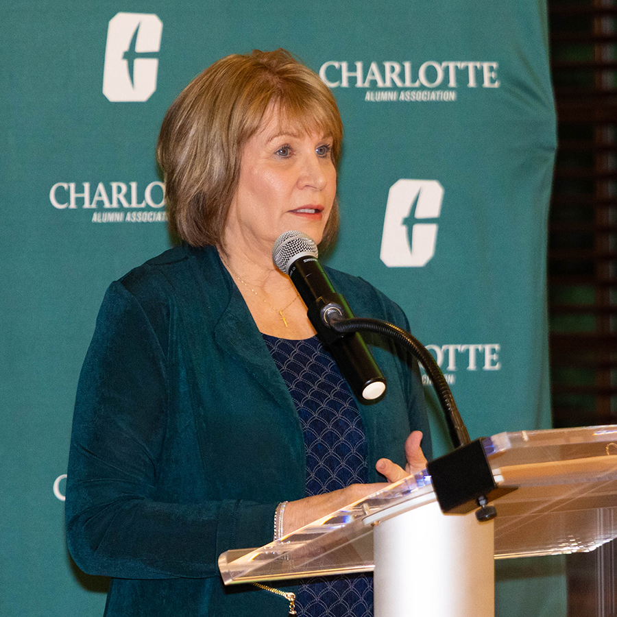 Jan Beatty-Hendley speaks at a podium during an event hosted by the Charlotte Alumni Association, with branding for the University of North Carolina Charlotte visible in the background.