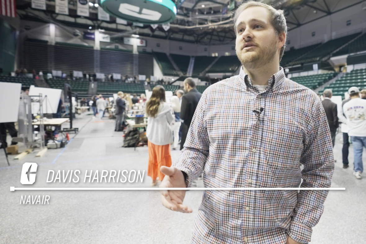 Davis Harrison of NAVAIR speaks during an interview inside a large arena hosting an engineering exhibition, with project displays and attendees visible behind him.