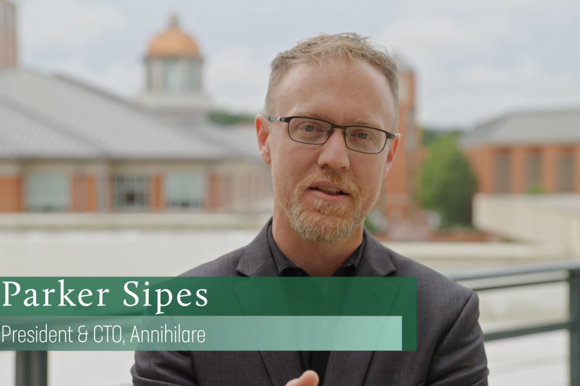 Parker Sipes, President and CTO of Annihilare, speaks on camera wearing glasses and a dark blazer, with UNC Charlotte campus buildings and a gold-domed tower in the background.